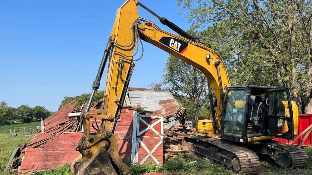 Yellow CAT excavator demolishing a red barn on a sunny day in rural farmland.