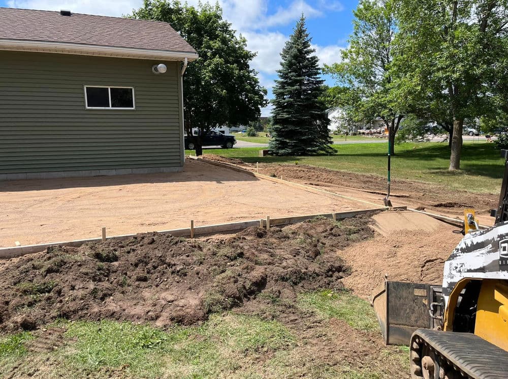 Construction site in progress with dirt, a house, and equipment for a new patio foundation.