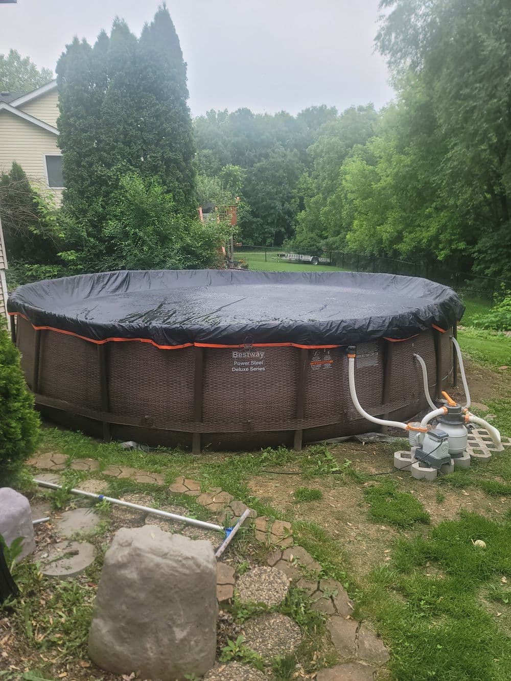 Round inflatable pool covered with a black tarp in a lush backyard setting.