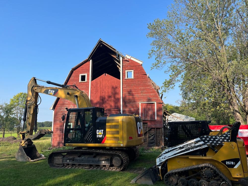 Excavator and skid steer next to a red barn under a clear blue sky.
