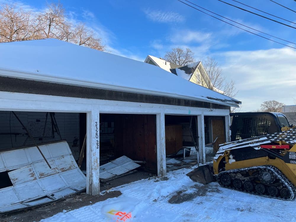 Damaged garage with snow on roof and construction equipment nearby under a clear blue sky.