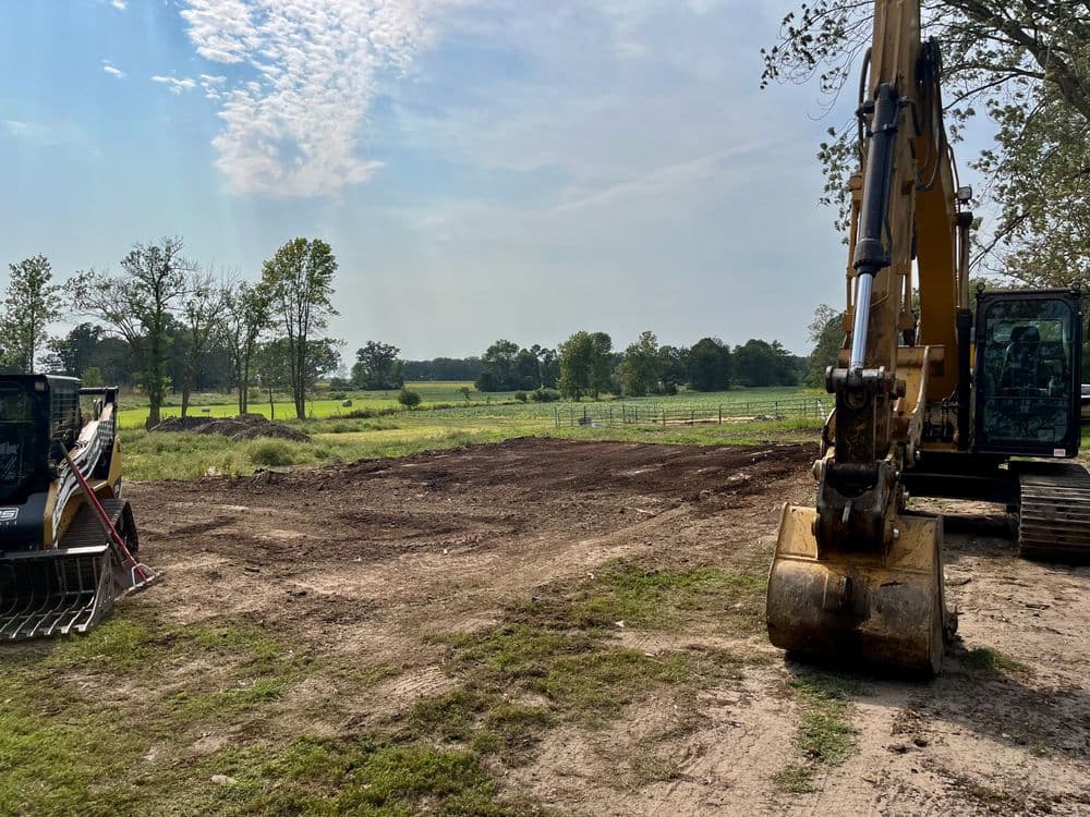 Construction equipment on a cleared rural site with trees and open fields in the background.