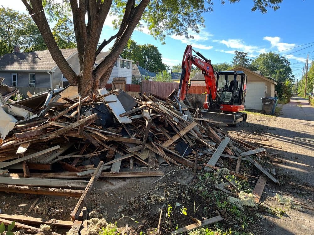Excavator near a pile of lumber debris in a residential area under a clear blue sky.