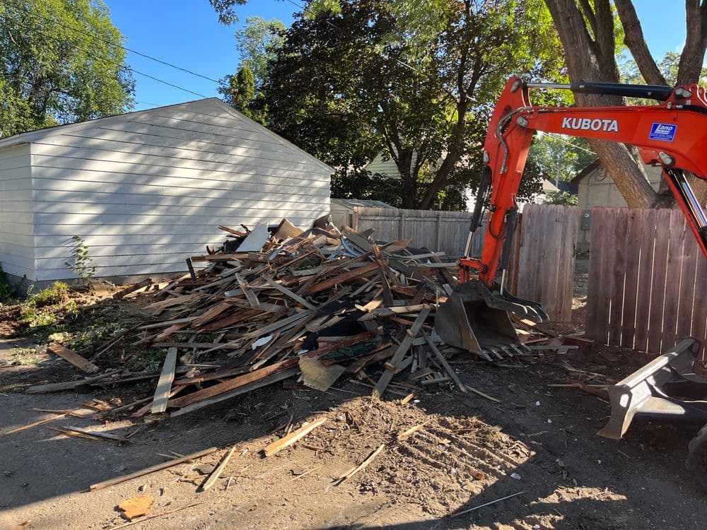 Excavator next to a large pile of debris behind a white house and wooden fence.