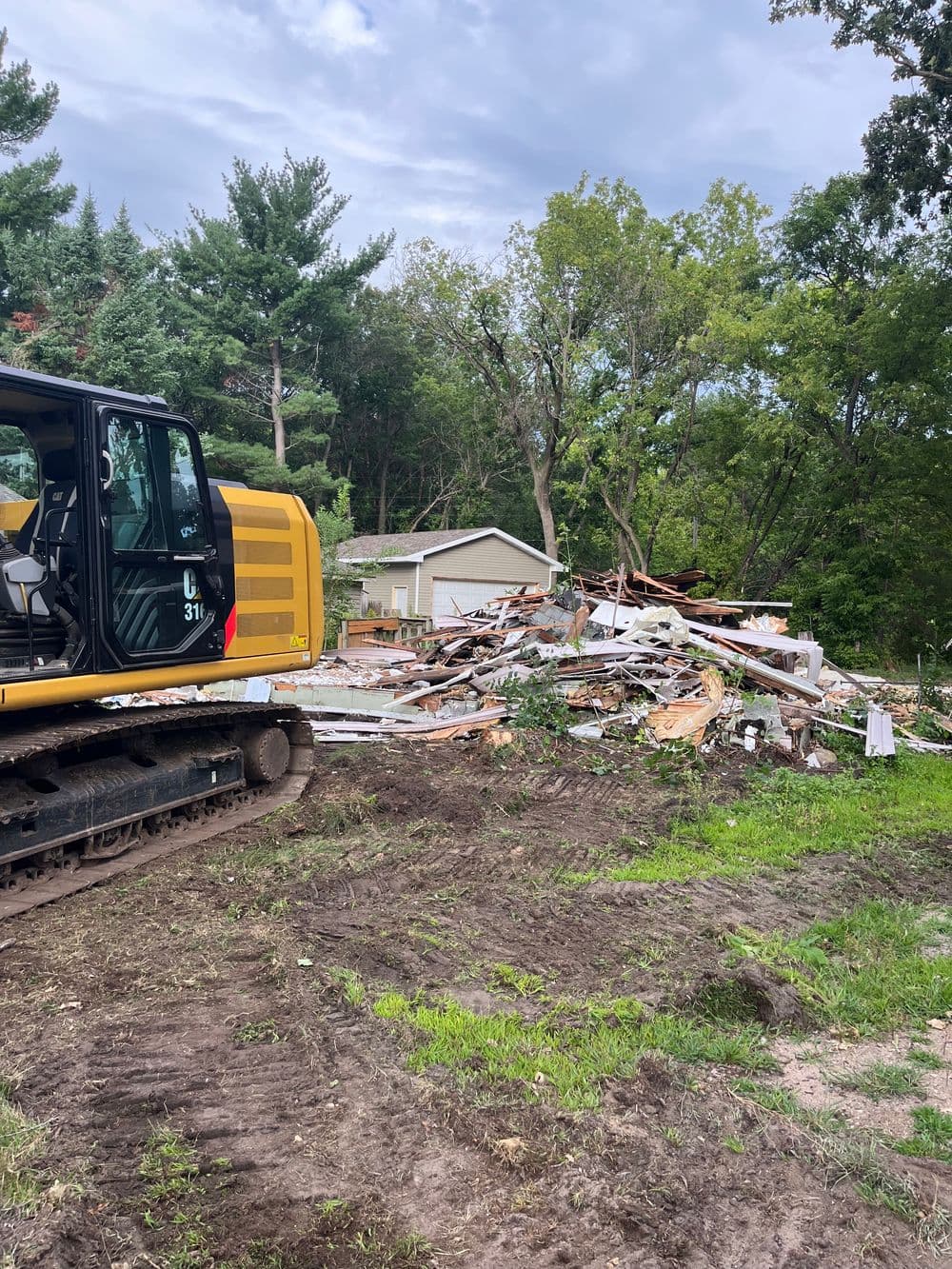 Excavator next to debris from demolished building in wooded area.