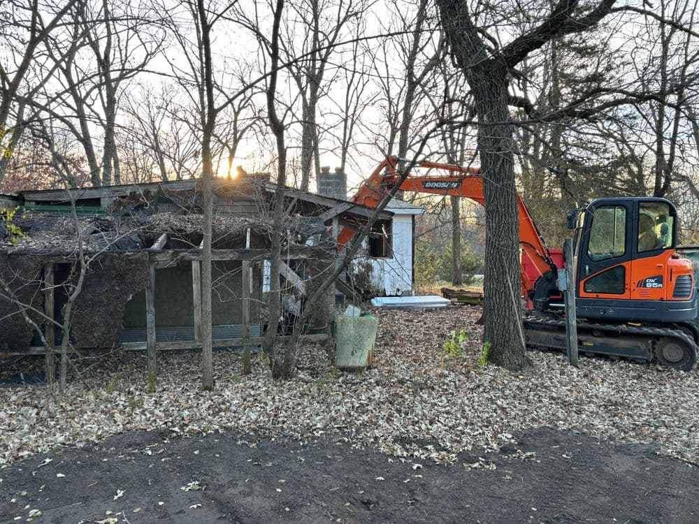 Excavator demolishing an old house surrounded by trees and fallen leaves at sunset.