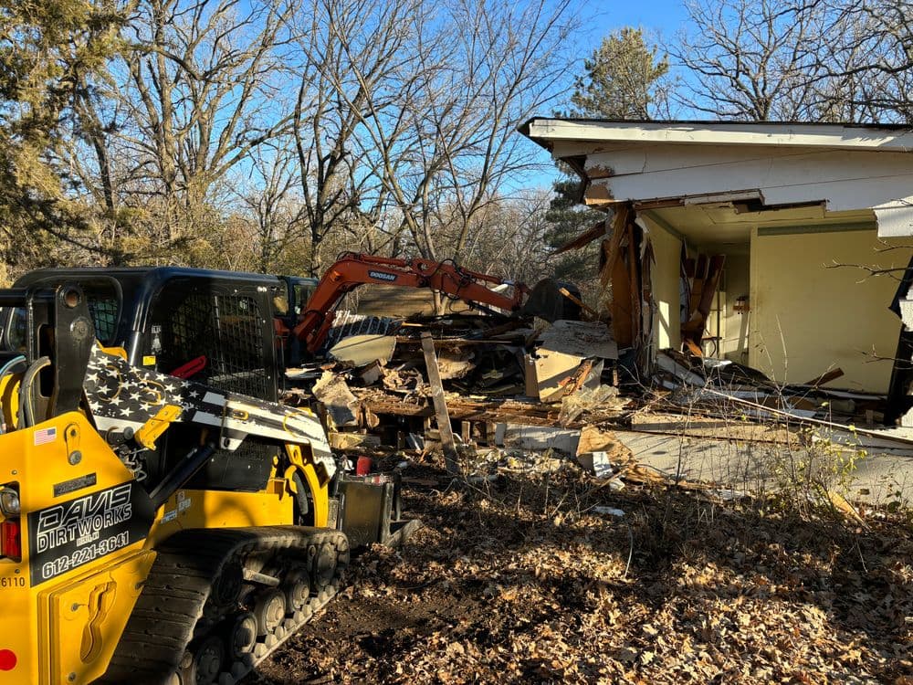 Excavators demolishing a building surrounded by trees and debris in a wooded area.